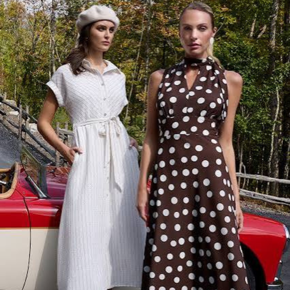 Two women standing next to a vintage car, one in a white dress and the other in a brown polka dot dress.